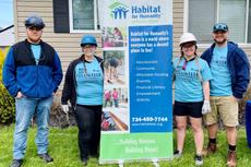 Four people in volunteer shirts, hard hats, and jeans stand by a Habitat for Humanity banner outside a house.