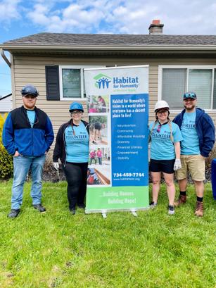 Four people in volunteer shirts, hard hats, and jeans stand by a Habitat for Humanity banner outside a house.