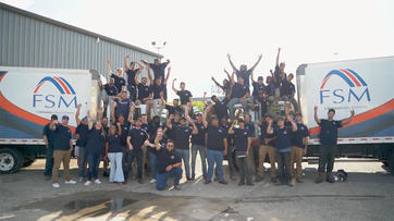 A large group of people in matching shirts pose and cheer between two FSM company trucks outdoors.