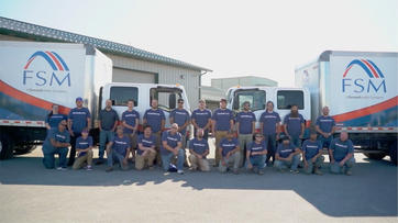A group of workers in matching FSM shirts pose in front of two FSM company trucks outside a building.