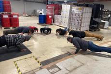 Seven people doing push-ups together on the floor of an industrial warehouse with pallets and barrels nearby.