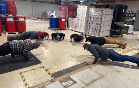 Seven people doing push-ups together on the floor of an industrial warehouse with pallets and barrels nearby.