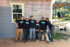 Six people in matching shirts pose and smile in front of a house under construction on a sunny day.
