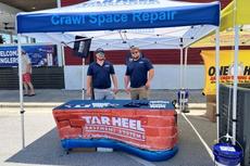 Two men stand at a Tar Heel Basement Systems booth under a tent labeled "Crawl Space Repair" at an outdoor event.