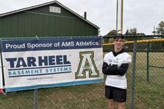 Teen in athletic gear stands by a fence with an AMS Athletics sponsor banner and school logos.