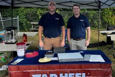 Two men stand behind a Tar Heel Basement Systems booth display at an outdoor event under a canopy.