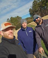 Three men in jackets and beanies pose for a selfie outdoors on a sunny day near a golf course.