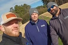Three men in jackets and beanies pose for a selfie outdoors on a sunny day near a golf course.
