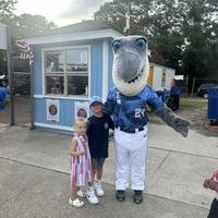 Two children pose with a person in a shark mascot costume outside a small blue building on a sunny day.
