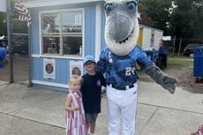 Two children pose with a person in a shark mascot costume outside a small blue building on a sunny day.