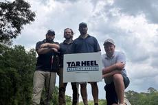 Four men pose on grass beside a "Tar Heel Basement Systems" sign under a cloudy sky.