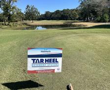 A "Tar Heel Basement Systems" sign stands on a golf course by a pond under a clear blue sky.
