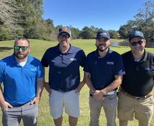 Four men in polo shirts and sunglasses stand on a sunny golf course with trees in the background.