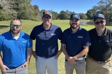 Four men in polo shirts and sunglasses stand on a sunny golf course with trees in the background.