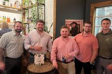 Five men in casual shirts stand around a barrel table in a rustic bar or restaurant, smiling at the camera.