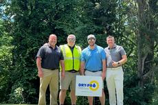 Four men pose on grass near trees with a "DRY-PRO" sign, dressed in golf and safety attire.