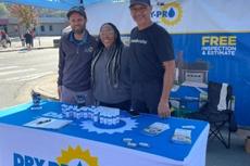 Three people stand smiling at a Dry Pro booth outdoors, with promotional items and a blue canopy overhead.