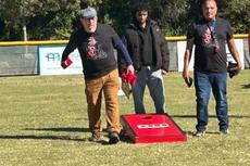 Three men play cornhole outdoors; one is tossing a bean bag toward a red cornhole board on grass.