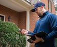 Groundworks CFI in blue jacket and cap uses handheld device and tablet for a foundation inspection outside a brick house near bushes.
