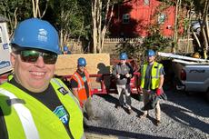 Four construction workers in safety gear smile at a building site with a red trailer and pickup truck nearby.