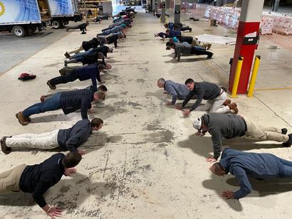 A group of people doing push-ups in rows inside a large industrial warehouse.