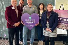 Five people smiling indoors, holding a heart-shaped "Unleashing Generosity" sign for Giving Tuesday.