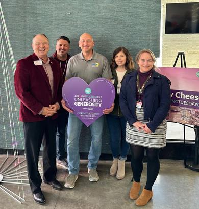 Five people smiling indoors, holding a heart-shaped "Unleashing Generosity" sign for Giving Tuesday.