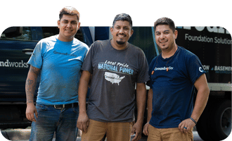 Three men in branded t-shirts stand smiling in front of a dark truck with "Groundworks" partially visible.