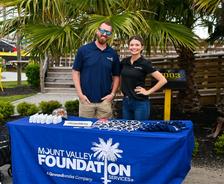 Two people stand behind a "Mount Valley Foundation Services" booth with sunglasses and items on the table.