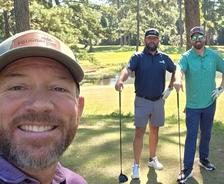 Three men smiling on a sunny golf course, two holding clubs, with trees and a pond in the background.