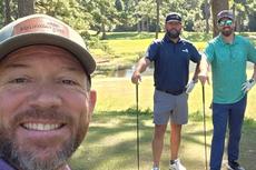 Three men smiling on a sunny golf course, two holding clubs, with trees and a pond in the background.