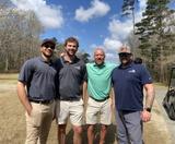 Four men in golf attire stand together outdoors on a sunny day, with trees and clouds in the background.