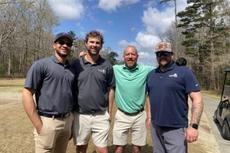 Four men in golf attire stand together outdoors on a sunny day, with trees and clouds in the background.