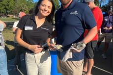 Two people smiling outdoors, holding oysters at a sunny event, with others gathered in the background.