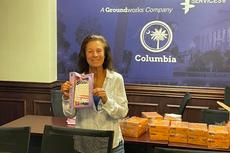 A woman smiles while holding a purple care package in an office with boxes stacked on a table behind her.