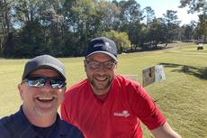 Two men in hats and sunglasses smiling on a sunny golf course with trees and signs in the background.