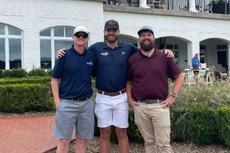 Three men in casual golf attire stand together smiling in front of a large white clubhouse building.