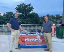 Two men stand by a Mount Valley Foundation Services table display outdoors in the evening.