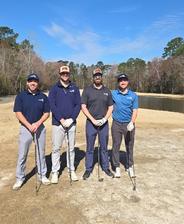 Four men stand outdoors on a golf course, each holding a golf club, with trees and a pond in the background.