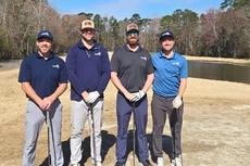 Four men stand outdoors on a golf course, each holding a golf club, with trees and a pond in the background.