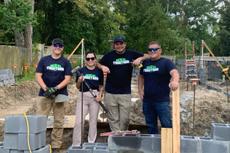 Four people in matching shirts stand smiling at a construction site with building materials and tools around them.