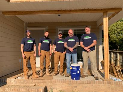 Five men in matching "Together" shirts stand on a porch under construction, posing for a group photo.