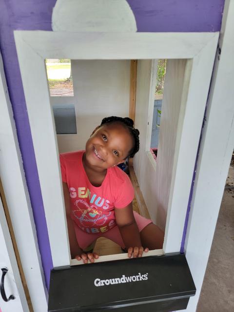Smiling girl in a pink shirt leans through a white playhouse window labeled "Groundworks.