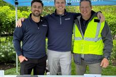 Three smiling men stand together under a blue canopy, one wearing a yellow safety vest, at an outdoor event.