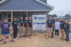 A group poses by a Habitat for Humanity sign in front of a newly-built house on a dirt lot.