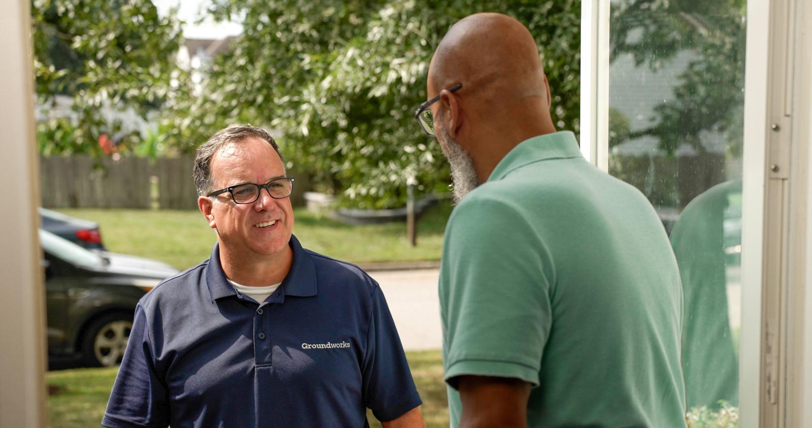 Two men talking at a doorway; one is outside wearing glasses and a blue shirt, the other is inside in a green shirt.