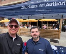 Two men stand and smile in front of an Ohio Basement Authority booth at an outdoor event with people seated in the background.