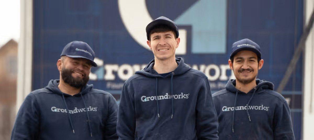 Three people wearing Groundworks hoodies and hats stand smiling in front of a large Groundworks sign.