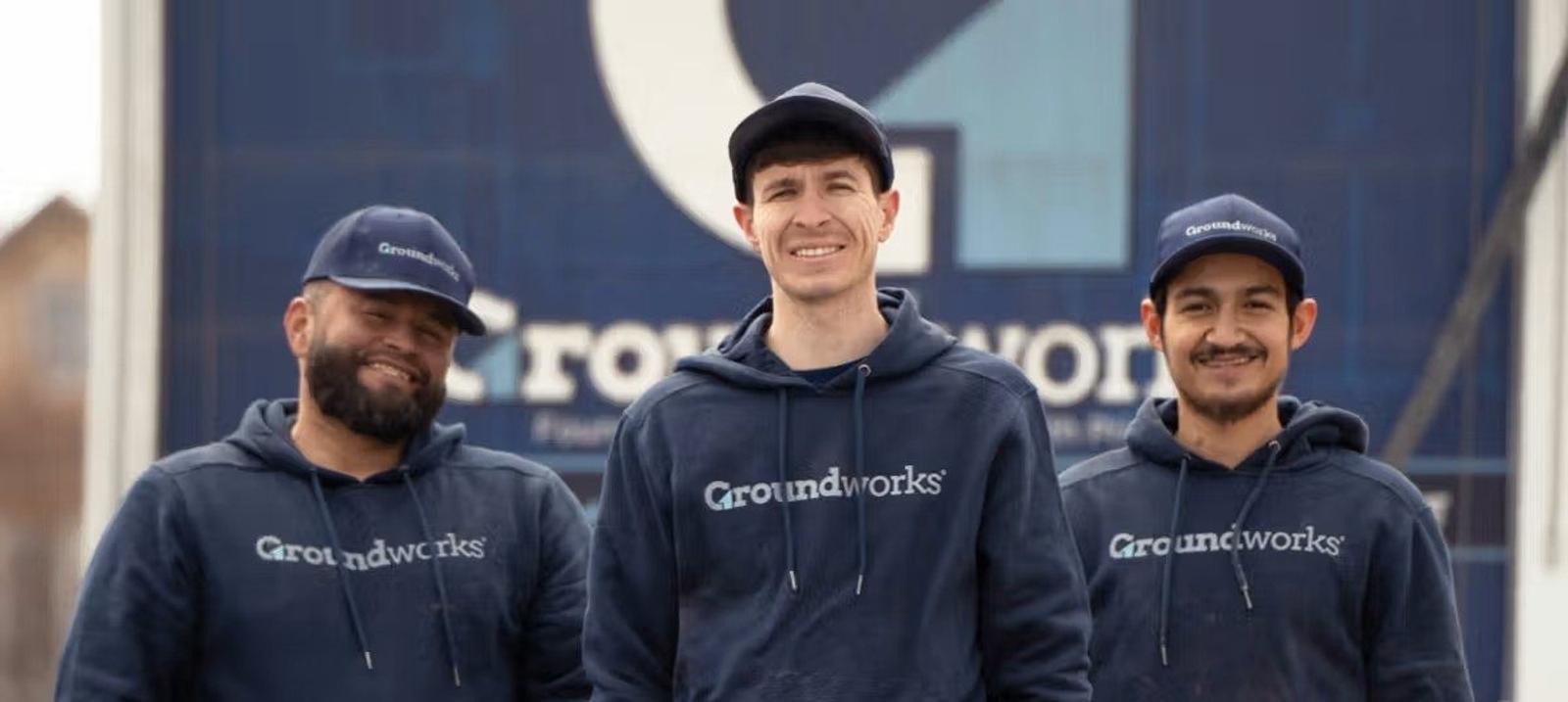 Three people wearing Groundworks hoodies and hats stand smiling in front of a large Groundworks sign.