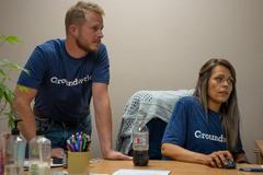 A man stands and a woman sits at a desk, both wearing “Groundwork” shirts, working in an office setting.
